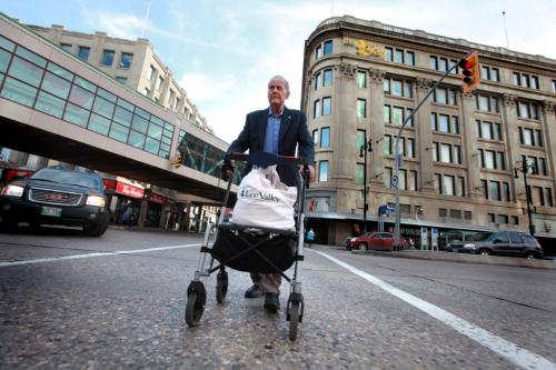 Eighty seven year old John Clark makes his way across Portage Ave. at Vaughan St. with his walker and small bag of groceries that he purchased at the grocery store in the basement at The Bay downtown.  Since he doesn't drive he is worried about where he will be able to buy his groceries once the grocery store closes. See Carol Sanders story. Oct 16,  2012 (Ruth Bonneville/Winnipeg Free Press)