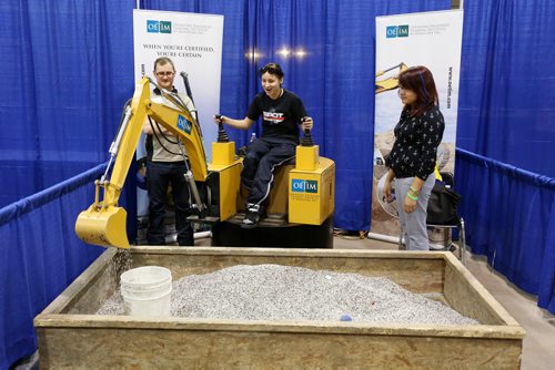 Brandon Sun 05032013 Fleming High School student Annie Cloud operates a scaled down digger as Billy Elias with the Operating Engineers Training Institute of Manitoba and Leigha Cloud of Maples Collegiate look on during the 28th Annual Brandon Career Symposium at the Keystone Centre on Tuesday. (Tim Smith/Brandon Sun)