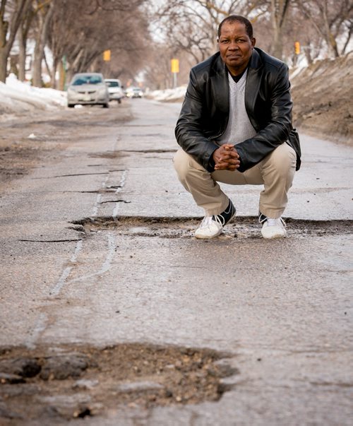 Richard Mutungi, owner of Winncity Driving School, stands in a large pothole on Inkster Blvd near Sinclair Street Thursday morning. Muntungi advises drivers to keep a safe distance, slow for potholes and don't swerve.  140313 - Thursday, {month name} 13, 2014 - (Melissa Tait / Winnipeg Free Press)
