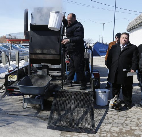 Pot Hole Repair -  Thunder Bird Restaurant, mayoral (right) candidate Mike Vogiatzakis in tie  in black dress overcoat  with (left) after pouring  rubber and asphalt  into  propane  heated  portable  heater Pellet Patch sales rep Saverio Marra director of sales demonstrates  pot hole reapair technology using asphalt and rubber     and  the effectiveness of a hot-mix-asphalt patching compound that had good results in Flint, Mich. Mar. 21 2014 / KEN GIGLIOTTI / WINNIPEG FREE PRESS