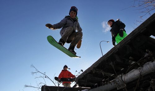 Duncan Maconachie, 12, takes a jump with his snowskate (skateboard sized snowboard) down a hill/embankment while his friends Noah van den Berg (left), 12, and Quinn Kinahan (right), 12, wait for their turn.  150111 January 11, 2015 Mike Deal / Winnipeg Free Press