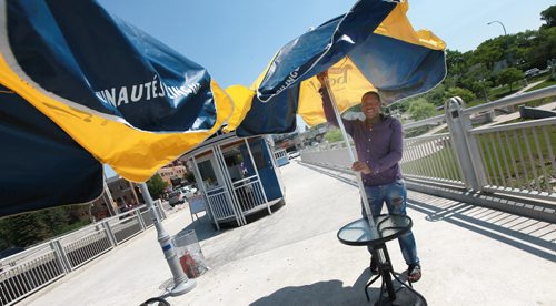 Serge Tamba opens up shade umbrellas at Regale de l'Afrique's kiosk, one of Three seasonal kiosks designed to look like horse-drawn trams from the early 1900s open each summer on the Plaza area at the east end of the Esplanade Riel bridge. Enterprises Riel, the economic development agency for the local French community, sets the kiosks up each year for the summer. Its tourism arm operates a tourist information centre out of one, and the the other two are food kiosks. One sells house-made frozen treats and the other features African cuisine. See Murray McNeil's story. June 26, 2015 - (Phil Hossack / Winnipeg Free Press)