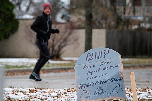 Daniel Crump / Winnipeg Free Press. A jogger reads cardboard tombstones, decked with protest slogans and the dates and ages of Manitoba COVID-19 victims, that have been setup outside the Premiers home on Wellington Crescent. October 31, 2020.