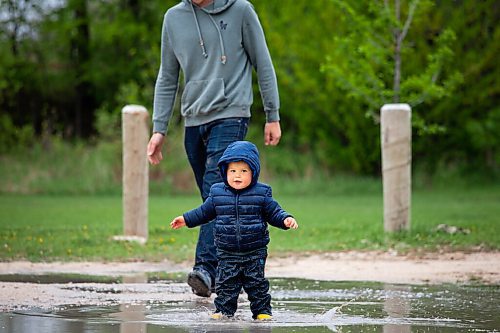 Daniel Crump / Winnipeg Free Press. Gil (right) Mcphail is watched by his dad Andrew McPhail as he splashes in a parking lot puddle near Sir John Franklin Park on a rainy Saturday afternoon. May 19, 2021.