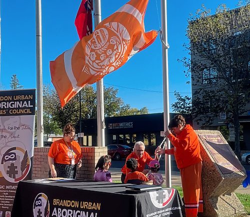 An orange and white Indigenous flag flaps gently in the breeze outside Brandon City Hall Friday morning, moments before it was raised by Deputy Mayor Glen Parker (kneeling), accompanied by several children from nearby daycares. Also helping were Shannon Saltarelli (left), the city's community housing and wellness co-ordinator, and Natashia Marion (right), Indigenous community co-ordinator with the Brandon Urban Aboriginal Peoples’ Council. (Michele McDougall/The Brandon Sun)