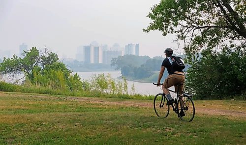 Wildfire smoke shrouds downtown Winnipeg high-rises as a cyclist makes their way along a bike path beside the Red River Friday morning. (Mike Deal/Winnipeg Free Press)